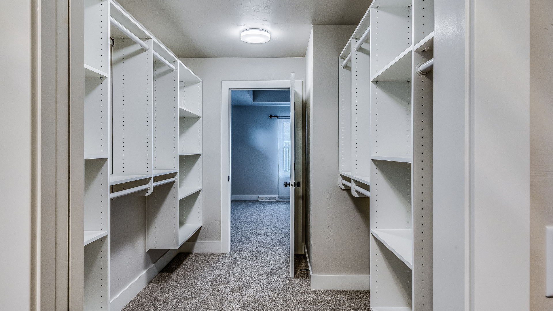 Narrow walk-through closet with white shelving, hanging rods, and plush carpeting leading to a bedroom.